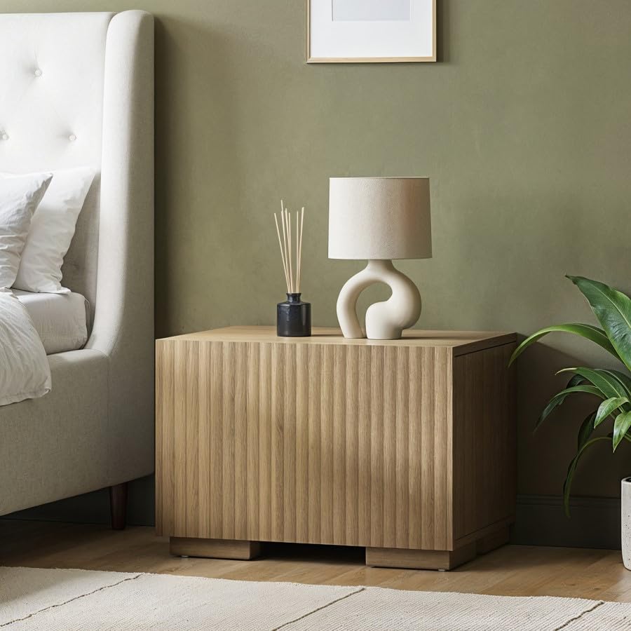 Wooden nightstand with decorative lamp and diffuser next to a bed in a bedroom.