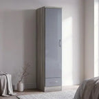 A tall, narrow wardrobe or storage cabinet with a light gray high-gloss door and a lower drawer, shown in a minimalist bedroom setting with a neutral wall, rug, and soft natural light.