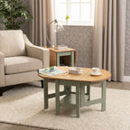 An oval coffee table with a light natural wood top and sage green painted legs, styled in a bright living room next to a beige armchair and a small wooden side table.