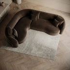 A curved, plush dark brown velvet sofa with matching throw pillows, styled on a light gray textured rug over a light wood herringbone floor.