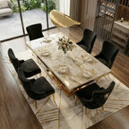 A modern rectangular dining table with a white marble-look top and gold base, surrounded by six black velvet upholstered chairs with gold trim, set on a geometric area rug in a luxury dining room.