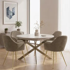 A round dining table with a white top and gold-tone X base, surrounded by four upholstered dining chairs in taupe with gold legs, set in a bright modern dining room.