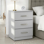 A three-drawer white nightstand with silver handles, styled next to a bed in a bright bedroom. A white table lamp 