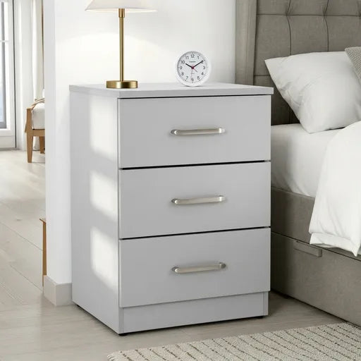 A three-drawer white nightstand with silver handles, styled next to a bed in a bright bedroom. A white table lamp 
