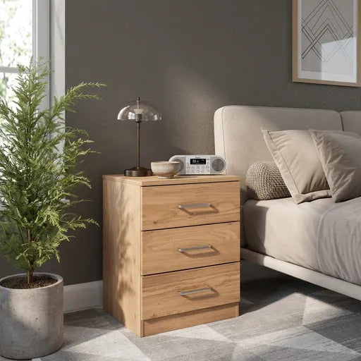 A three-drawer wooden nightstand with a light oak finish stands beside a bed in a modern bedroom. 