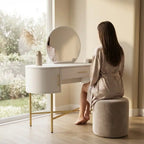 Woman sitting at a vanity table in a bright room with large windows.