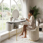 Woman sitting at a white vanity table in a bright room with large windows.