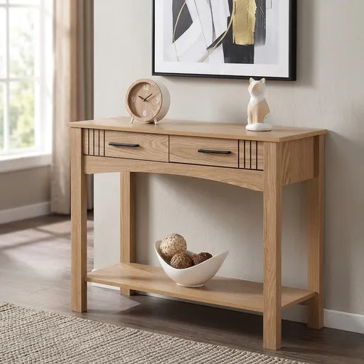 A light oak wood console table with two drawers and an open lower shelf, styled in a bright room with a clock, ceramic cat figurine, and bowl of decorative balls on top.