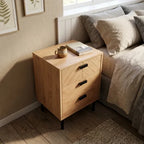 A light oak wood nightstand with three drawers, black geometric handles, and black legs, styled next to a bed in a bedroom setting.